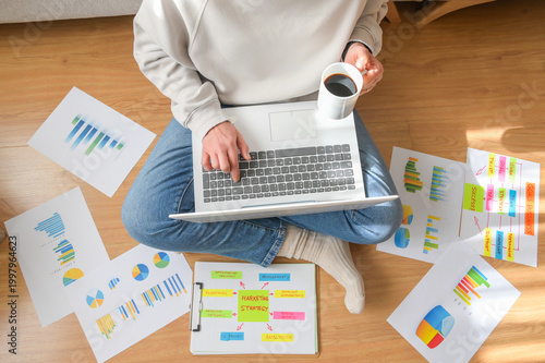 Photos Person sitting on a wooden floor using a laptop to work remotely, surrounded by