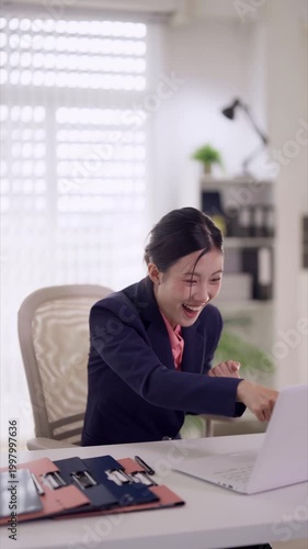 excited Asian businesswoman celebrating success achievement raise fists winner gesture sitting desk laptop computer modern office expressing joy excitement