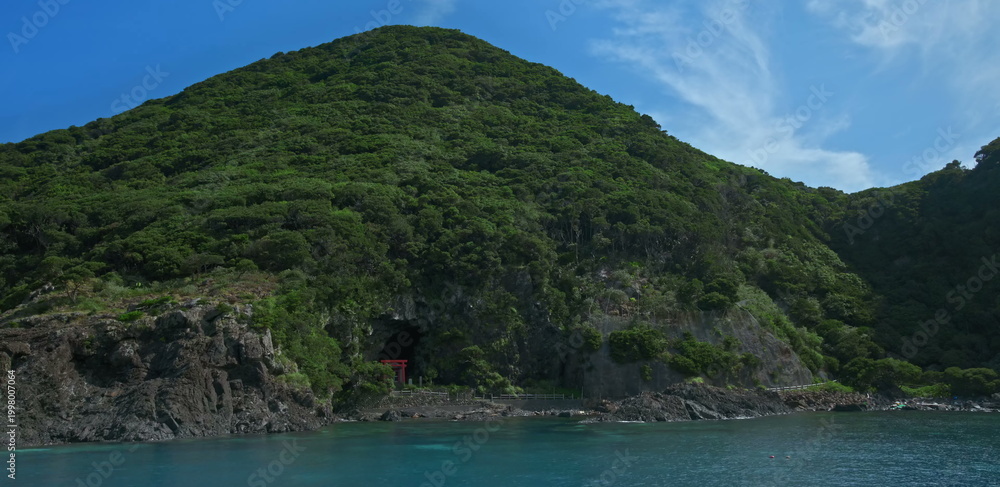 Fototapeta premium Coastal sea cave with red torii gate and lush green mountain slopes