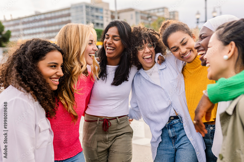 Obraz premium Diverse young women hugging and smiling together outdoors. Cheerful group of friends expressing unity and affection naturally. Friendship and joy concept.