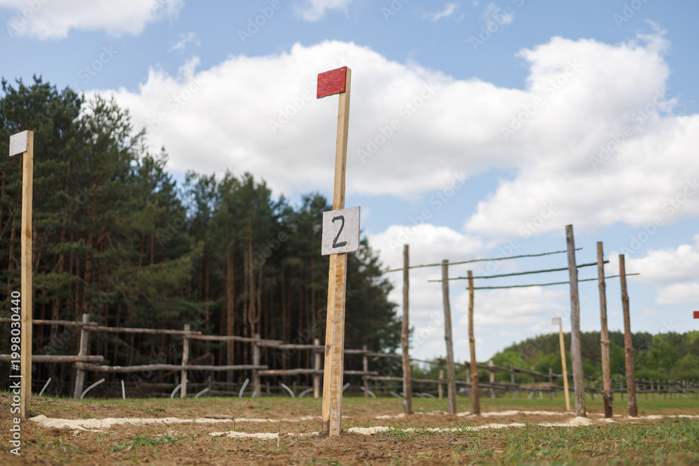 Fototapeta premium Wooden post with number 2 sign and red flag on an outdoor track. White powder marks the path on the grass. Forest and blue sky background