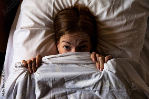 Foto A woman lies in her bed, pulling the white blanket up to her eyes