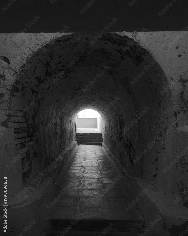 Obraz premium Moody stone tunnel with wet floor leading toward bright exit and stairs, symbolizing hope, transition, or escape.