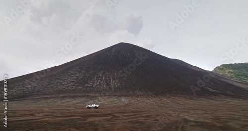 Pick-up truck crossing a dark volcanic ash plain at the base of Mount Yasur on Tanna Island, Vanuatu, under cloudy sky, remote off-road adventure near an active volcano. Tracking side view shot