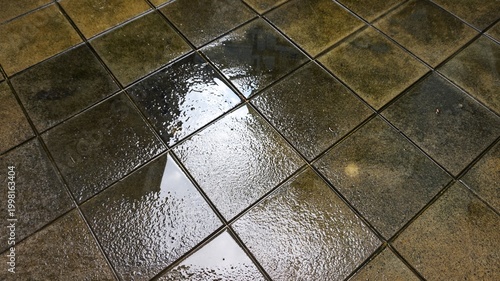wet ceramic floor with puddles of water. Stone-like square tiles arranged in a grid pattern create a textured background.