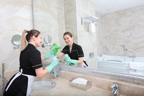Maid in uniform wipes bathroom mirror. Girl smiles, looking at camera. Marble countertop. Cleaning at home or at hotel. Hands in protective gloves. Concept of cleanliness and order