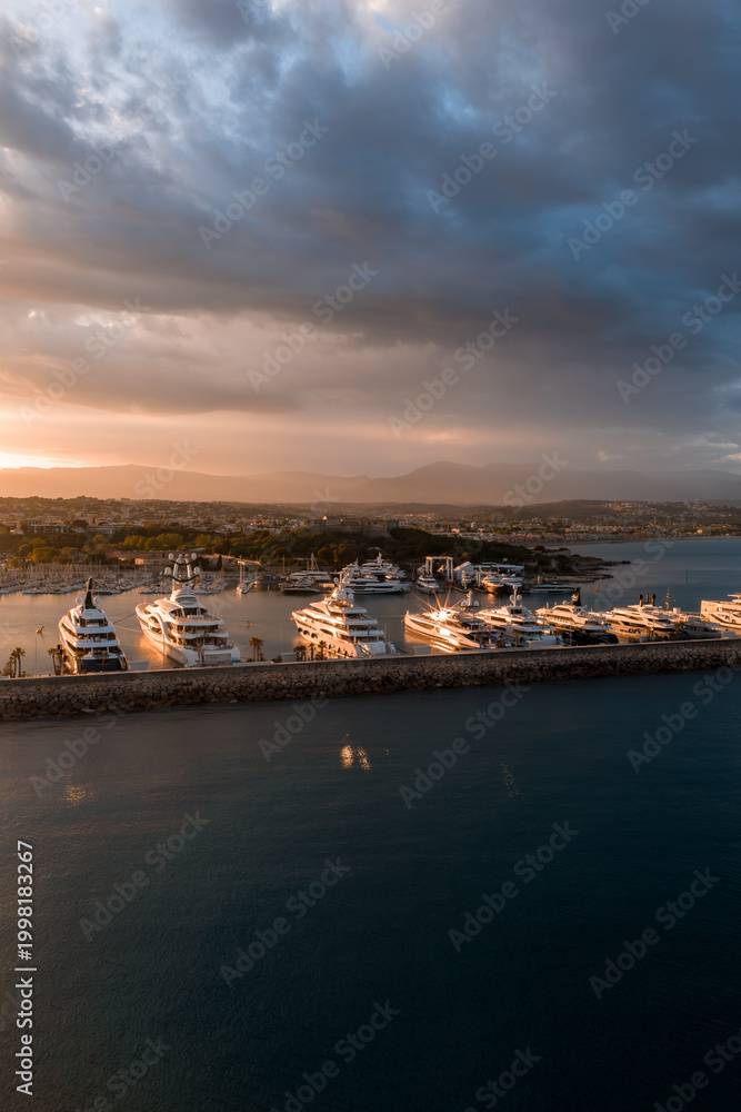 Fototapeta premium Aerial view of Port Vauban in Antibes, France, with rows of superyachts along the breakwater at sunset. The harbor curves to the old town, with Alps faintly visible.