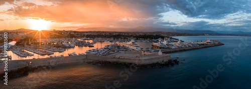 Aerial view of Port Vauban in Antibes at sunset shows old town, Fort Carre, quays with yachts and superyachts, warm light on hulls, curving breakwaters, calm water.