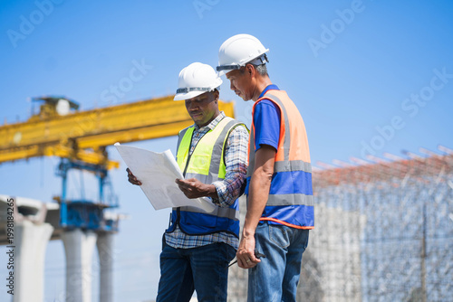 African American and asian engineers coordinating infrastructure work with radio, diverse engineers discussing a bridge construction project with blueprints on site