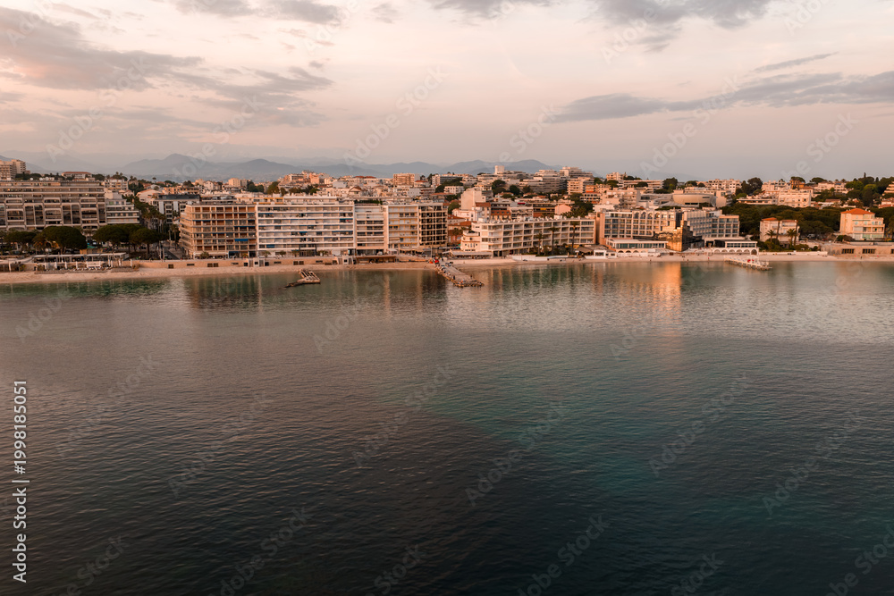 Fototapeta premium Aerial view of Antibes, France, at sunset shows modern mid rise hotels and apartments by a calm bay, old town cluster, sandy beaches, pastel clouds, and distant hills.