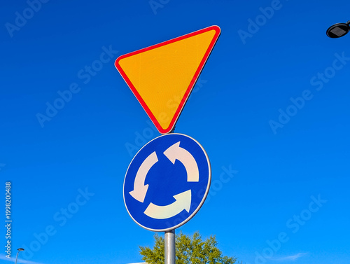 Warsaw, Poland, March 01, 2026. Road signs including a roundabout and a yield triangle sign under a bright sunny sky. High quality photograph.