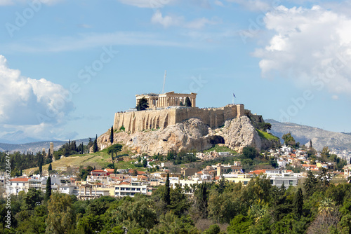 Photography Panoramic view of the ancient Acropolis of Athens with the Parthenon temple on top, surrounded by lush green trees and traditional Greek architecture under a blue cloudy sky
