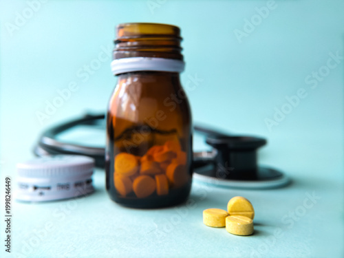 Amber glass bottle filled with round, yellow pills, partially open on a light blue surface. A stethoscope is slightly blurred in the background, suggesting a medical setting. 