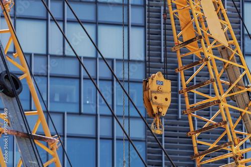 Fotografie Yellow crane boom with 50-ton crane block and hook suspended by steel cables towers above urban development site adjacent to modern glass‑paneled building visible in background