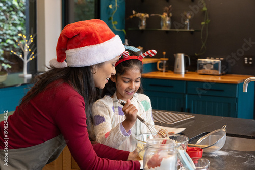 Indian female mother-daughter with Santa hat and antlers mixing batter at kitchen island with whisk