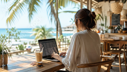 Woman working on laptop at tropical beachside cafe, enjoying refreshing drink and ocean view
