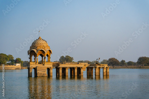 Traditional sandstone pavilion standing in calm waters of Gadisar Lake with reflection and clear sky in Jaisalmer.