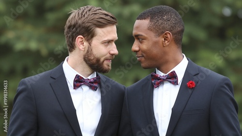 Diverse same-sex couple grooms looking at each other during outdoor marriage ceremony