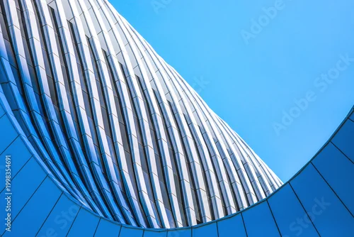 Obraz Modern building facade featuring dynamic architectural curves and flowing lines under a bright blue sky, creating an abstract geometric pattern with sleek steel and glass panels