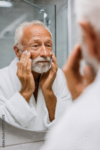 Senior man applying moisturizer cream on face looking in mirror, skincare routine for elderly male, healthy aging, grooming in white bathrobe, daily self care, lifestyle