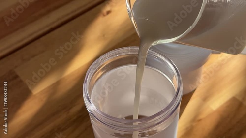 White liquid pours from a pitcher into a transparent plastic tumbler on a wooden surface in warm sunlight, filling the cup smoothly.