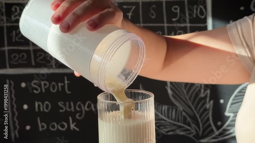 Person pours a protein shake from a blender bottle into a ribbed glass against a chalkboard with healthy lifestyle goals.