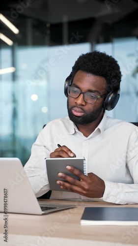 African american businessman in headphones watching video call conference takes notes looking at laptop screen sitting at workplace in business office. Worker listening online training. Vertical video