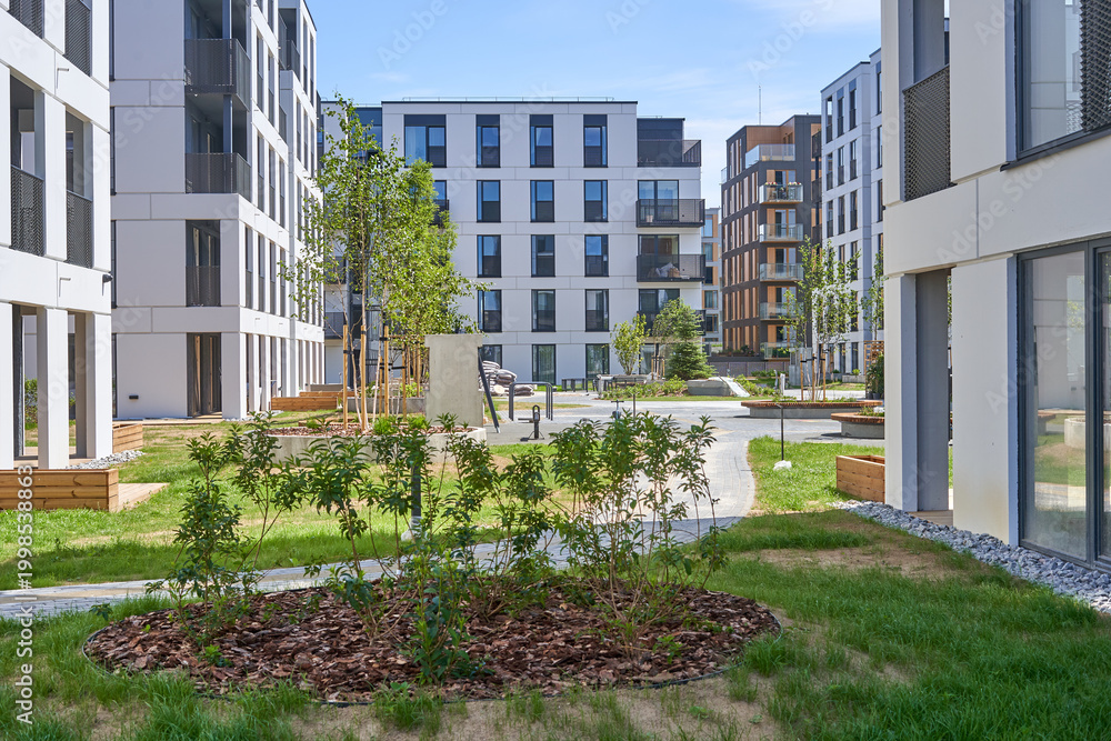 Fototapeta premium A bright, sunlit view of a modern residential courtyard surrounded by contemporary apartment buildings. The architecture features clean, minimalist white facades with large dark-framed windows and