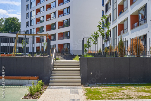 Modern residential apartment complex featuring multi-story buildings with white facades and orange-accented balconies. A central wide concrete staircase with metal handrails leads upward between