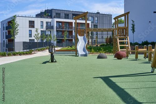 Photography A modern outdoor playground featuring wooden play structures with a silver wavy slide, monkey bars, and climbing equipment on a green rubberized safety surface