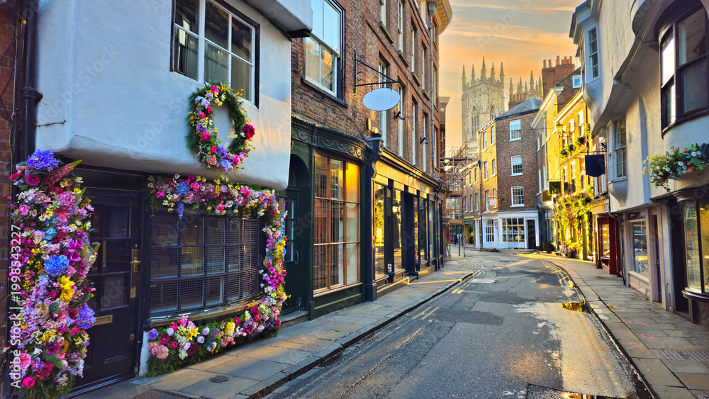 Fototapeta premium View through the Old Town streets of York, England at sunset with flower lined shop facades and York Minster in the background