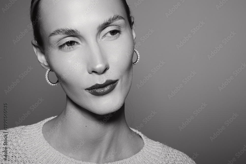 Fototapeta premium Close-up portrait of a slender Caucasian woman with light skin and short hair wearing earrings and a knitted sweater, looking sideways in a monochrome studio setting.