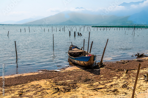 Photography A view of a sampan boat beached beside an oyster farm on the Lap An Lagoon in Vi