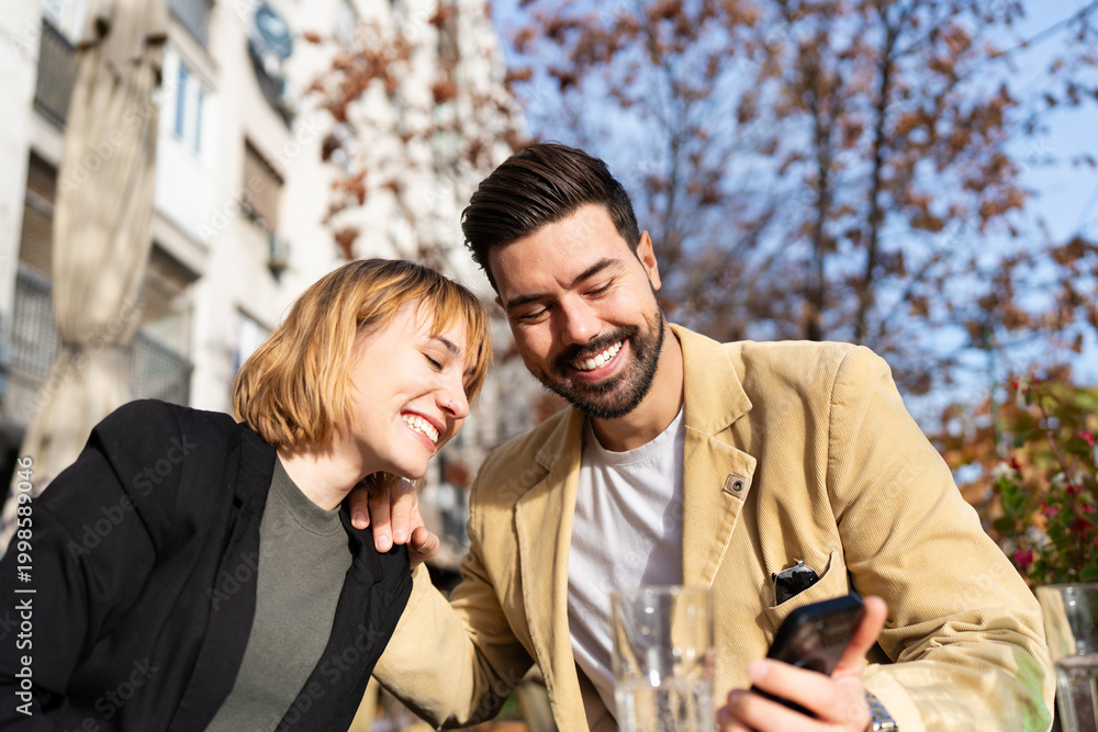 Fototapeta premium A happy young man and woman enjoy a sunny afternoon together at an outdoor city cafe, laughing while looking at content on a smartphone.
