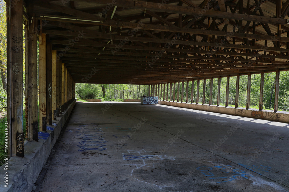 Fototapeta premium Lange, leerstehende Holzscheune mit offener Balkenstruktur auf dem Bienitz, skelettartiges Holzgebäude im Wald bei Sonnenlicht, Burghausen, Leipzig, Sachsen, Deutschland