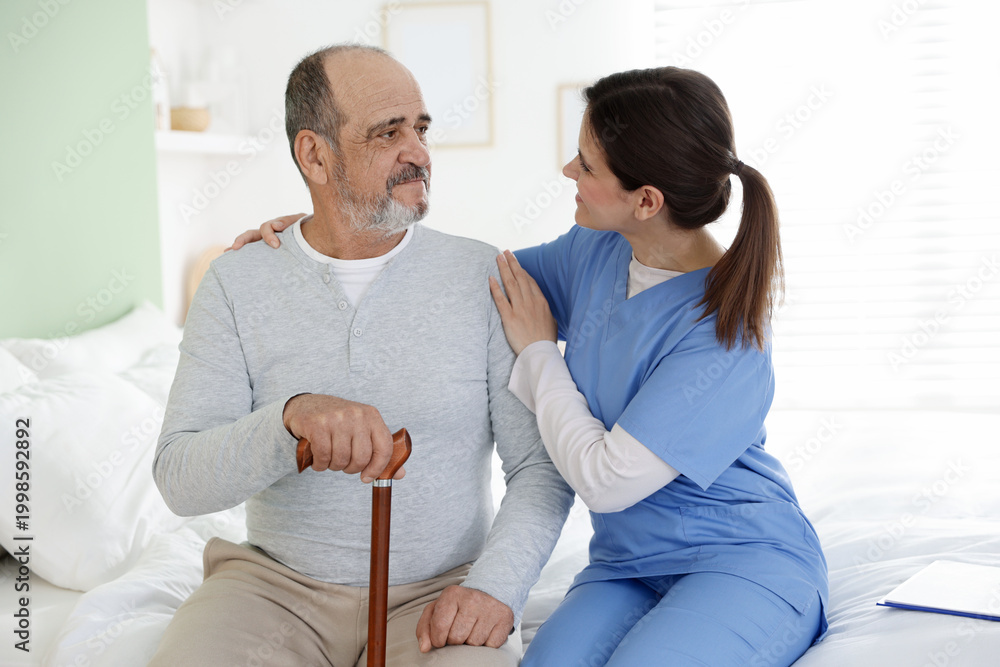 Fototapeta premium Care worker supporting senior man with walking cane on bed at nursing home