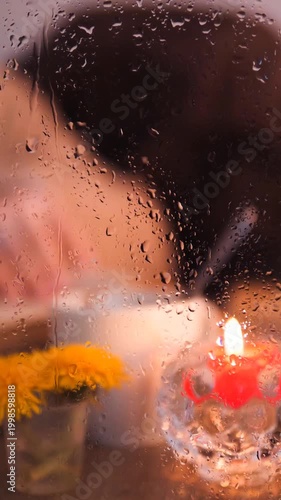 Stirring Coffee and Turning Book Pages Behind Rainy Window, Cozy Interior Scene with Candlelight and Yellow Flowers