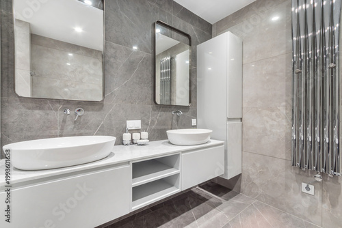 The space shows a modern bathroom layout featuring two sinks on a white counter. Large mirrors hang above, and light reflects on tiled walls. A towel warmer is installed beside storage cabinets
