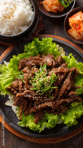 A steaming plate of Korean bulgogi, thinly sliced marinated beef glistening with caramelized sauce, garnished with sesame seeds and scallions, served on a bed of crisp lettuce leaves with a side of ki