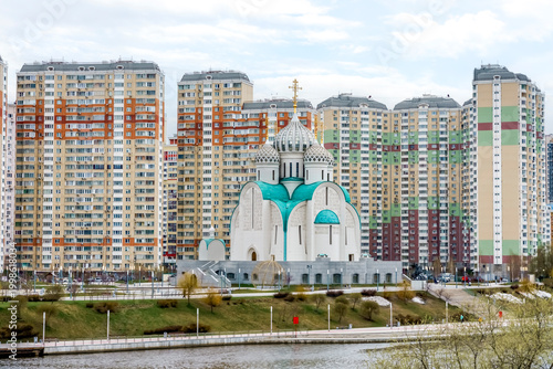 Modern Orthodox church with green domes situated between high-rise residential buildings along a riverbank in a contemporary urban landscape