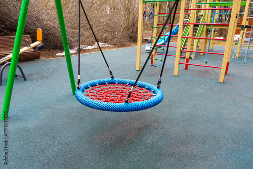 Circular swing with blue and red rope design hanging from green frame in a playground setting, surrounded by climbing structures and benches