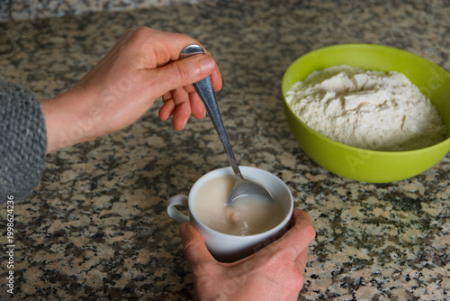 Manos de mujer revuelven con una cuchara de metal levadura de panadería en una taza con agua, con un cuenco con harina dentro de fondo