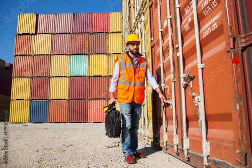 Man in yellow hard hat and orange safety vest standing beside stacked shipping containers at a port