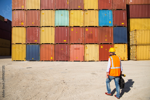 Man in yellow hard hat and orange safety vest standing beside stacked shipping containers at a port