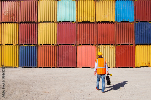Man in yellow hard hat and orange safety vest standing beside stacked shipping containers at a port