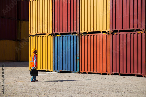Man in yellow hard hat and orange safety vest standing beside stacked shipping containers at a port