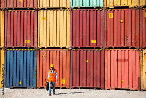 Man in yellow hard hat and orange safety vest standing beside stacked shipping containers at a port