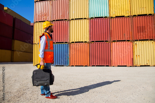 Man in yellow hard hat and orange safety vest standing beside stacked shipping containers at a port