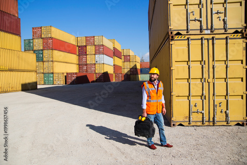 Man in yellow hard hat and orange safety vest standing beside stacked shipping containers at a port