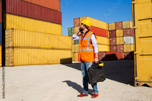 Man in yellow hard hat and orange safety vest standing beside stacked shipping containers at a port